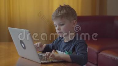 金发的学龄前男孩在笔记本电脑上打字，微笑着看着电脑屏幕。 孩子在家坐在网上学习