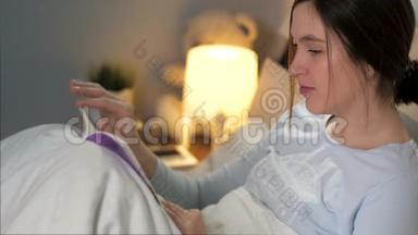 女孩在看书。 年轻迷人的白种人女人晚上躺在床上，拿着毯子看书和喝酒
