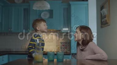 金发的学龄前儿童在厨房里和妈妈说话，桌上放着苹果汁。 孩子给妈妈讲故事。