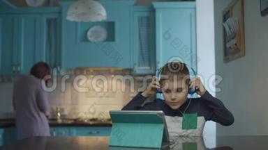 自闭症男孩在厨房里使用带耳机的平板电脑，而母亲则带零食。 患有自闭症的孩子