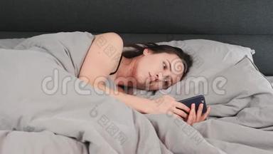 年轻女孩躺在灰色的床上，在智能手机上打字的肖像。 布鲁内特女士躺在空床上，看着智能手机。 魅力