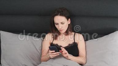 愤怒的女人躺在床上在智能手机屏幕上打字。 深色头发的女孩子在灰色的床上躺着，用触摸屏打电话