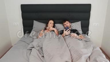 女人和男人躺在床上，手里拿着智能手机。 年轻的已婚夫妇在灰色的床上看着设备，在网络上发短信