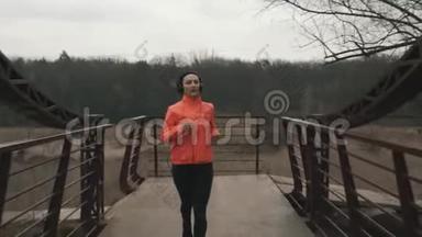 布鲁内特的女运动员戴着耳机跑过公园的人行桥。 穿橙色夹克的年轻女子在桥上跑步