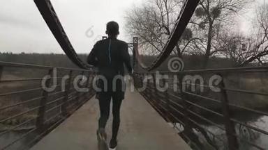 瘦小的男人在公园的桥上横渡河，背面的景色。 男运动员在户外跑步。 穿黑色紧身衣的年轻人
