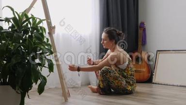年轻漂亮的女人坐在家里的地板上，拿着画笔，在画布上画画。 艺术学院或绘画学校。 激励
