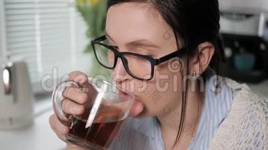 女孩喝绿茶或<strong>红茶</strong>。 厨房里戴着眼镜的漂亮年轻女人的特写镜头慢慢地喝着棕色的<strong>饮料</strong>