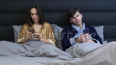 已<strong>婚</strong>夫妇在床上看智能<strong>手机</strong>。 家庭夫妇使用设备躺在床上，科技上瘾的夫妇。 女人和男人