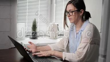 自由职业者的工作理念。 漂亮的带眼镜的年轻女人在厨房工作，看手机和笔记本电脑