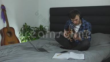 专业吉他老师在电吉他上练习新和弦.. 戴耳机的人在家里的床上独奏吉他。 青年