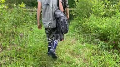 母亲和儿子带着背包的游客，徒步旅行者沿着长满了高草和高草的小路走到河边