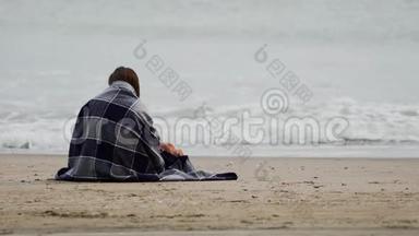 风和日丽的春日，坐在海边的布鲁内特女孩裹着温暖的格子毯子。 大海上有波浪。 后视图