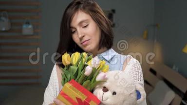 女孩带着<strong>礼物</strong>，迷人的年轻女子独自在房间里，手里拿着一束<strong>鲜花</strong>，<strong>礼物</strong>，软玩具，看着。