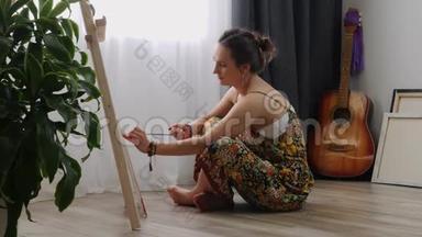 女艺术家在画布上画画，用丙烯酸颜料坐在家里的地板上。 年轻害羞的女孩用刷子画图片。 内部资料