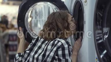 一个年轻的积极的女人穿着格子衬衫，在家用<strong>电器商店</strong>里选择洗衣机。 往里面看