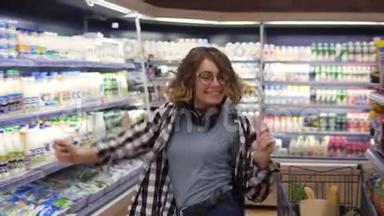 在超市：快乐的年轻女孩在超市的货架之间滑稽地跳舞。 穿牛仔裤和黑色的卷发女孩