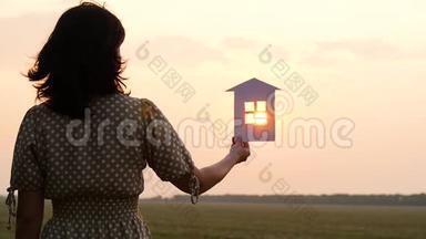 女孩手里拿着纸房子的模型。 日落时太阳的光芒穿过太阳的窗户