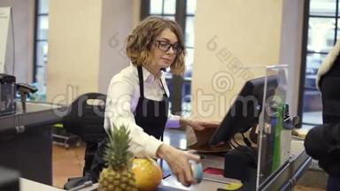 穿着白色衬衫和黑色围裙扫描产品的开朗苗条的女售货员，在明亮的<strong>收银台</strong>买水果