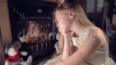 一个小悲伤失望的女孩坐在人造壁炉旁，在柔软的玩具旁边感到悲伤。 被宠坏的概念