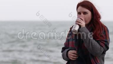小女孩拿着一个金属<strong>热</strong>水瓶，在靠近大海的河堤上放着<strong>一杯热饮料</strong>。 外面大风，头发长在
