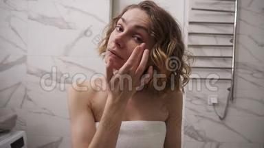 一位身穿白色大毛巾的年轻女士正在<strong>眼部</strong>下面的面部涂奶油，按摩动作，看镜子