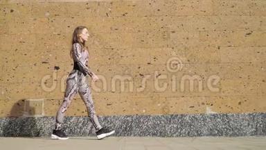 穿着<strong>运动服</strong>的漂亮年轻运动<strong>女孩</strong>优雅地沿着棕色的混凝土墙走。 慢动作。 对斯泰迪达姆的射击