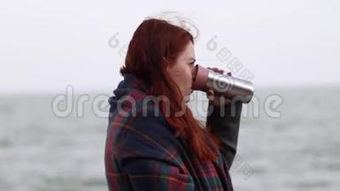 年轻的红头发女孩拿着一个金属<strong>热</strong>水瓶，在靠近大海的河堤上放着<strong>一杯热饮料</strong>