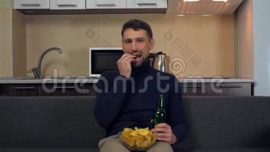 一个穿着深蓝色套衫和灰色裤子的年轻人坐在沙发上，吃<strong>薯片</strong>，喝啤酒，看电视上的娱乐节目