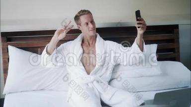 穿着白色浴袍的金发帅哥在床上用手机拍自拍照片