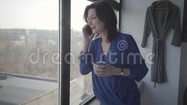 带<strong>着</strong>咖啡杯微笑的白种人女人的肖像，<strong>看着窗外</strong>，在电话里交谈。 自信快乐