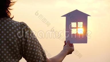 一个女孩手里拿着纸房子模型的剪影。 日落时太阳的光线穿过