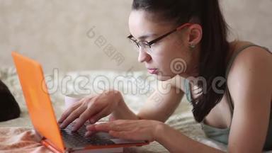 年轻漂亮的女孩正在用她的笔记本电脑工作，躺在家里的床上。 一个年轻可爱的女孩躺在床上打字