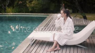 穿着白色浴袍的女人坐在靠近游泳池的日光浴床上，清晨喝咖啡