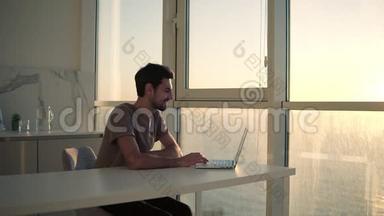欧洲深色头发的男人坐在工作室厨房的全景窗口附近-看着笔记本电脑桌面，兴高采烈