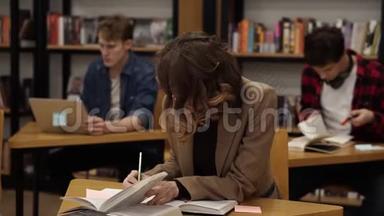 一个穿着棕色夹克的古丽的欧洲女孩坐在大学<strong>图书馆</strong>里写论文，准备考试，然后看上去