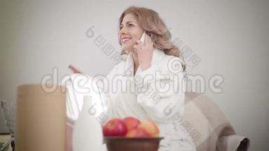穿着白色浴袍的开朗的白种人女孩坐在沙发上打电话的肖像。 美丽的女人