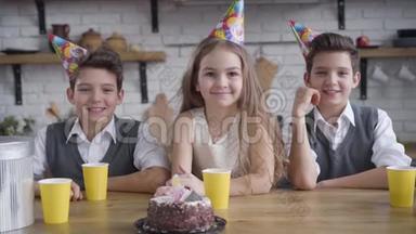 慢动作，两个白种人<strong>双胞胎</strong>男孩和戴着派对帽的<strong>可爱</strong>女孩端着生日蛋糕坐在桌子旁，看着