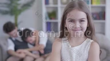 快乐的小黑发女孩在镜头前微笑，<strong>跑到</strong>双胞胎兄弟在沙发上玩的背景。 肖像