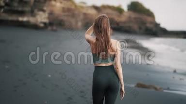 后景，身穿运动服的女士沿着黑沙滩散步，转身