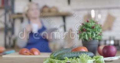 厨房的餐桌上摆着新鲜蔬菜的特写镜头，模糊的女人一边喝着咖啡，一边在厨房看<strong>菜谱</strong>
