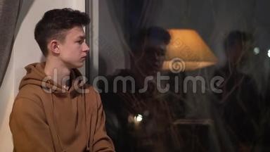 侧观年轻的黑发白种人男孩坐着看着窗外。 令人不安的男孩独自度过一个晚上。 悲伤