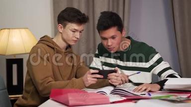 两个现代的多种族朋友坐在桌子旁，使用智能手机。 微笑青少年大学生
