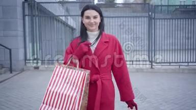 中镜头前看到穿红色外套的优雅年轻女士带着购物袋在城市里散步。 积极的高加索妇女