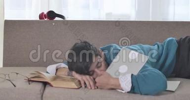 迷离的白人成年男孩躺在沙发上看书和写字板。累了的男大学生在室内睡着了