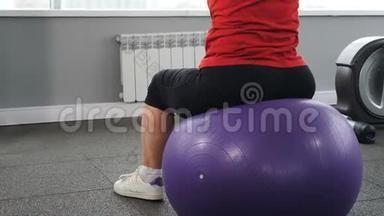退休妇女坐在健身球上健身房锻炼的背面。 积极和健康的养老金领取者的概念。 长者