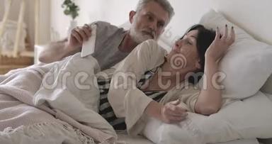愤怒的高加索老人在床上拿走妻子`智能手机。 丈夫愤怒地看着黑发女人在里面聊天