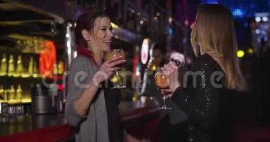 两个年轻漂亮的女人在夜总会的酒吧柜台旁用鸡尾酒碰杯跳舞。 积极女孩