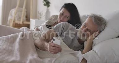 可疑的高白种人妻子看着丈夫的肩膀在智能手机屏幕。 成熟男人在网上聊天