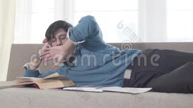 精疲力竭的白人年轻人看书打哈欠。 戴眼镜的成年大学生睡着了