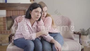 沮丧的白种人母女坐在家里的沙发上。 少女抱着妈妈，成年女子抱着头